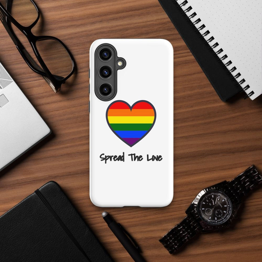 a white phone case with a rainbow heart and the text "Spread The Love" on a wooden surface, surrounded by various office and personal items such as a laptop, glasses, a notebook, a pen, and a watch.