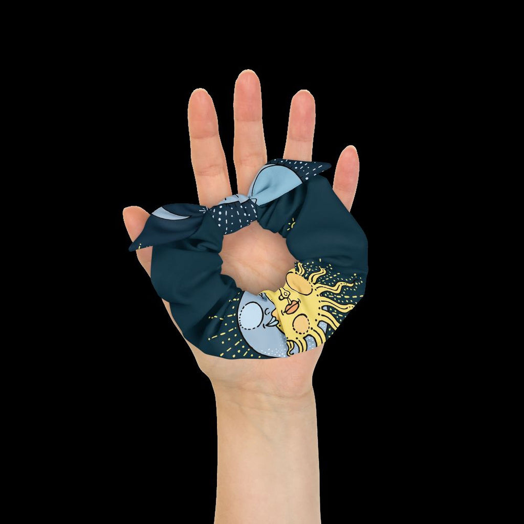 A hand holding a blue and yellow scrunchie with a sun and moon design against a black background.