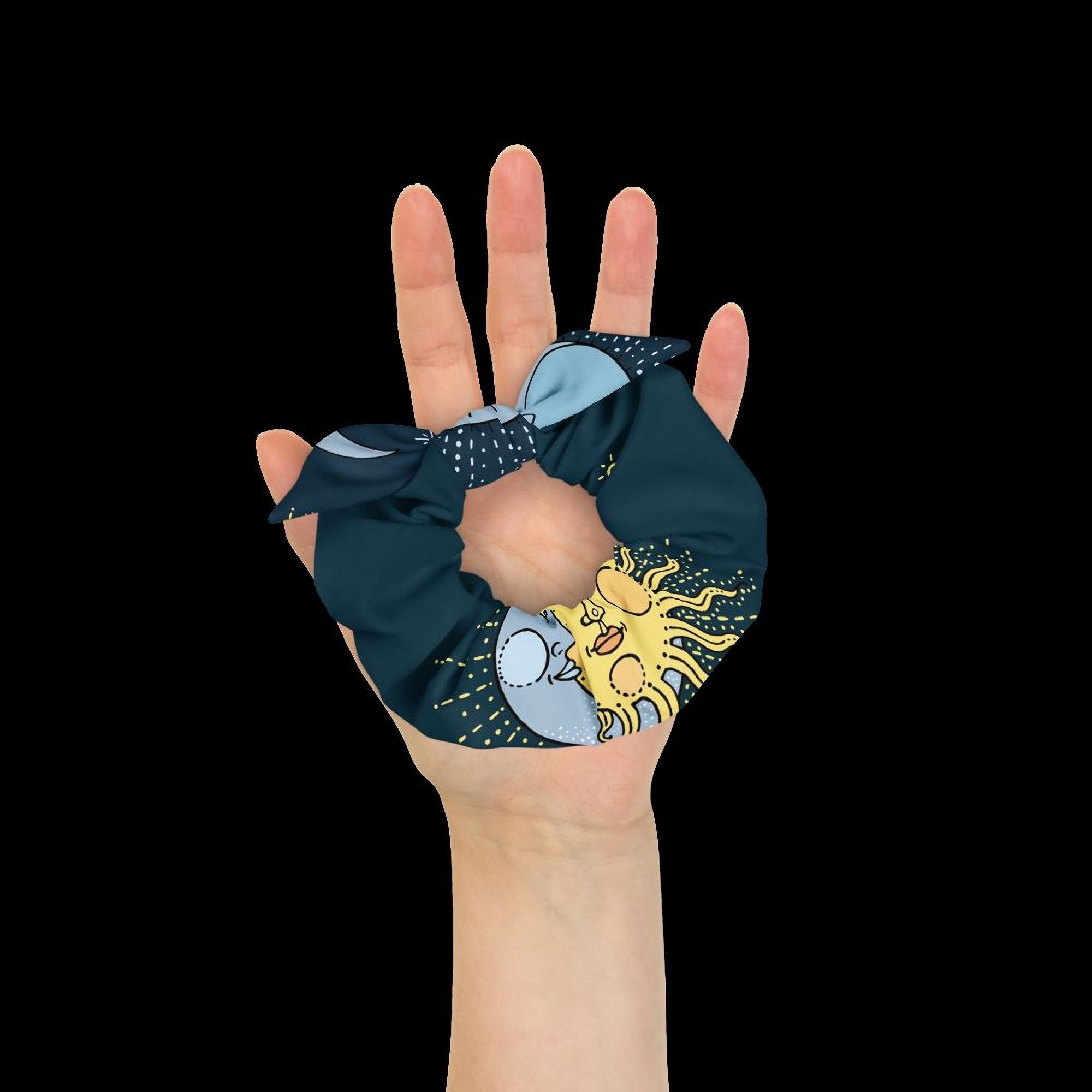 A hand holding a blue and yellow scrunchie with a sun and moon design against a black background.
